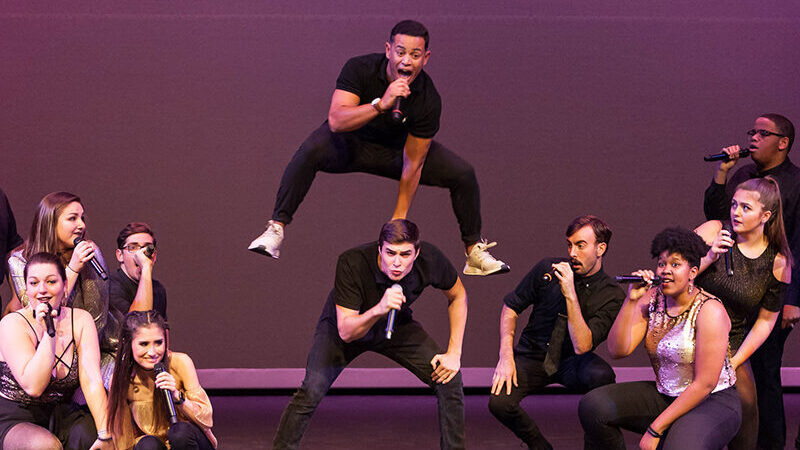 Image of a group of a cappella performers singing on stage. One of them is vaulting over the back of another.