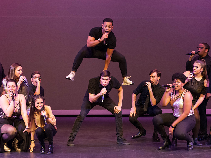 Image of a group of a cappella performers singing on stage. One of them is vaulting over the back of another.