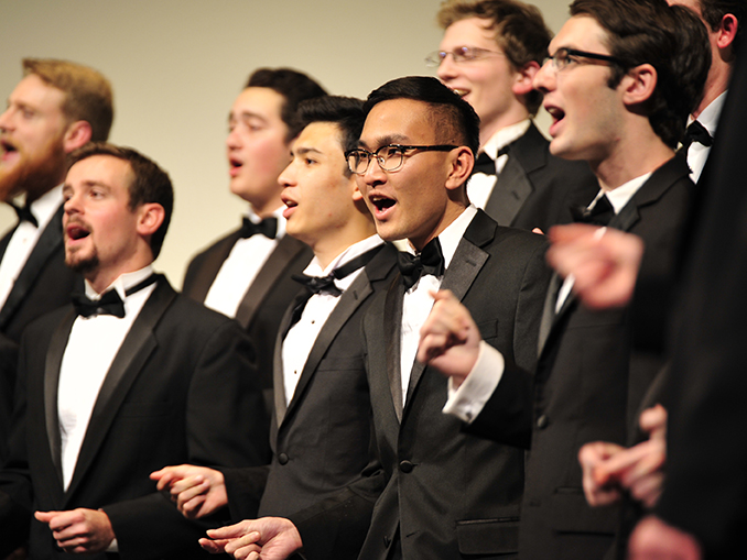 Photo of a group of male singers performing with the choir wearing tuxedos and snapping their fingers.