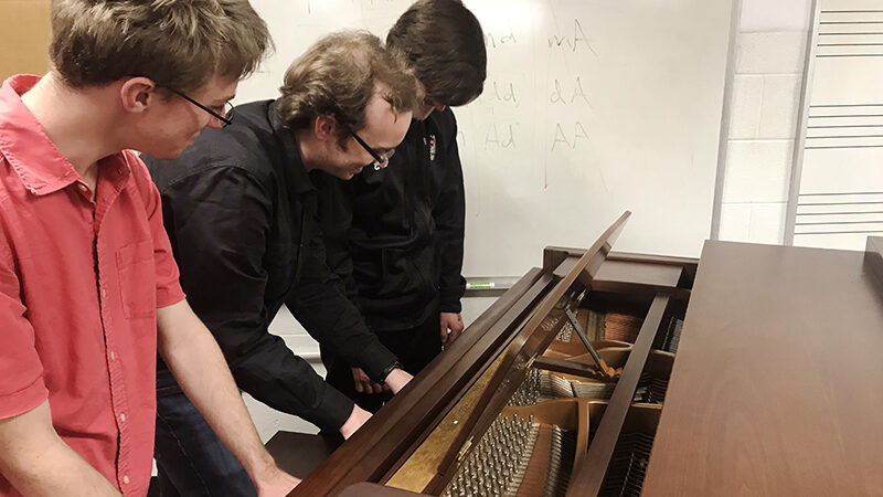 Three students stand looking down at a piano keyboard together.