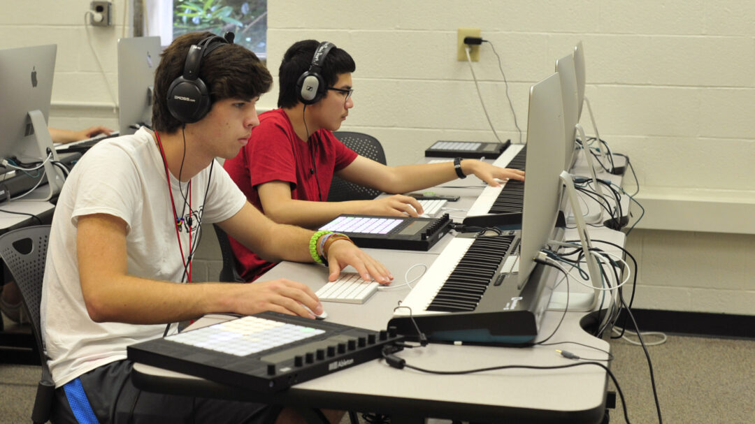 Students wearing headphones work on electronic music compositions in a computer lab equipped with music keyboards and midi controllers.