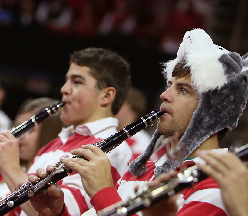 Clarinet players in the Varsity Pep Band perform during a basketball game wearing the striped pep band shirts. One player wears a hat in the shape of a wolf head.
