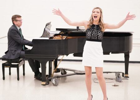 A female student sings with her arms raised during a music minor recital. Behind her, a man accompanies her on the piano.