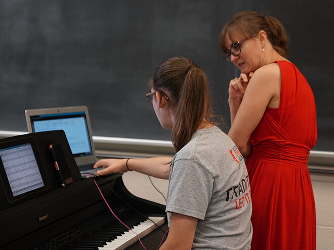 A student seated at a digital keyboard shows Dr. Olga Kleiankina something on her tablet screen.