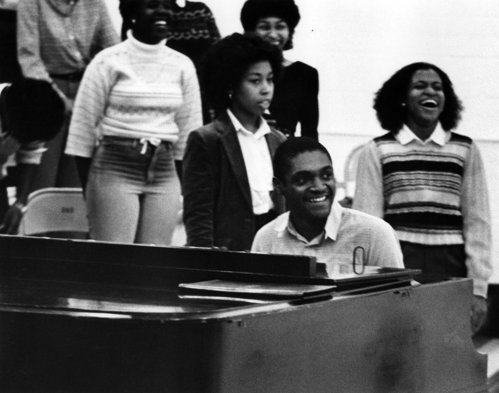 Image of a student sitting at a piano with fellow members of New Horizons Gospel Choir on risers behind him.