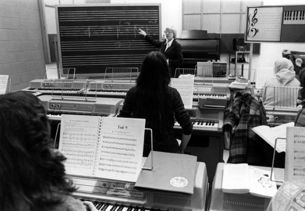 Black and white image from the 1970s of Phyllis Vogel teaching at a blackboard while students sit at digital pianos.