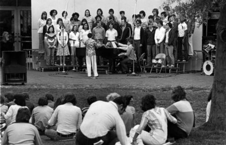 Image of a choir on risers outdoors, performing for students seated on the grass.