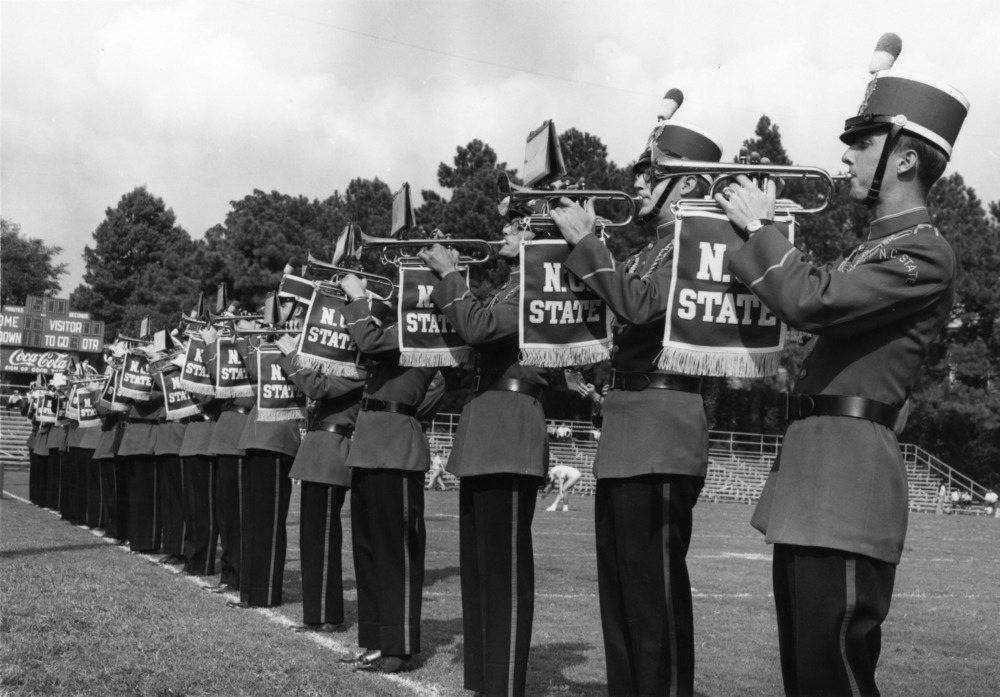A line of marching band trumpeters play their instruments in a black and white photo from the 1960s.
