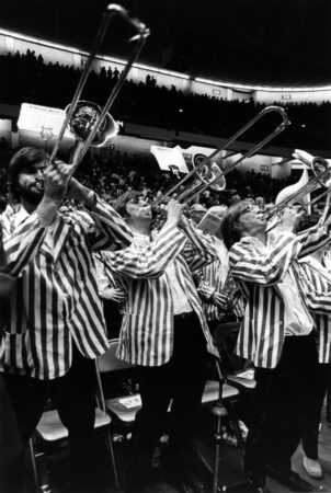 Three trombone players in striped jackets perform during a basketball game with the pep band in a black and white photo from the 1970s.