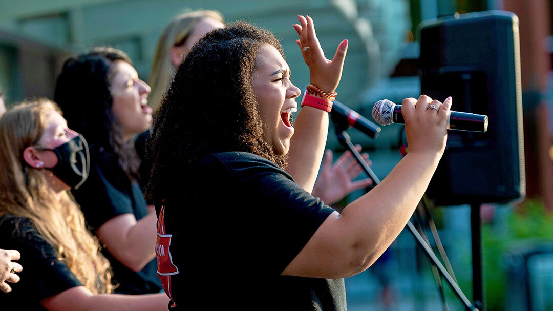 A student sings into a microphone outdoors, with other members of Chordination a cappella singing in the background.