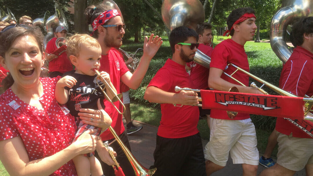 Eva Feucht holds her young son who is holding a toy trombone, while the marching band trombone section walks past them.