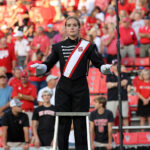 Laura Dugan stands on a ladder in uniform with her hands raised. A crowd of fans wearing red and white is behind her in the stands.