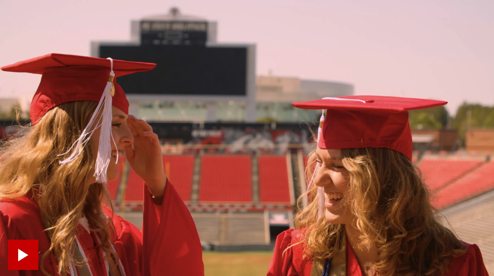 Screenshot of Alyssa and Emily wearing their caps and gowns with the field and seats of Carter-Finley Stadium in the background.