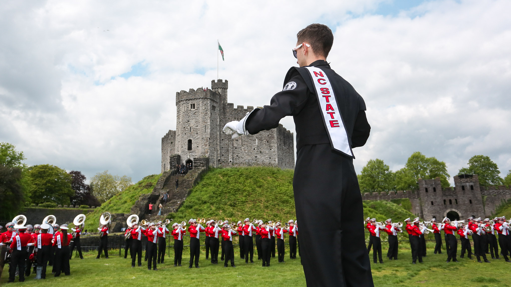A student drum major leads their marching band section through a performance in front of a European castle.