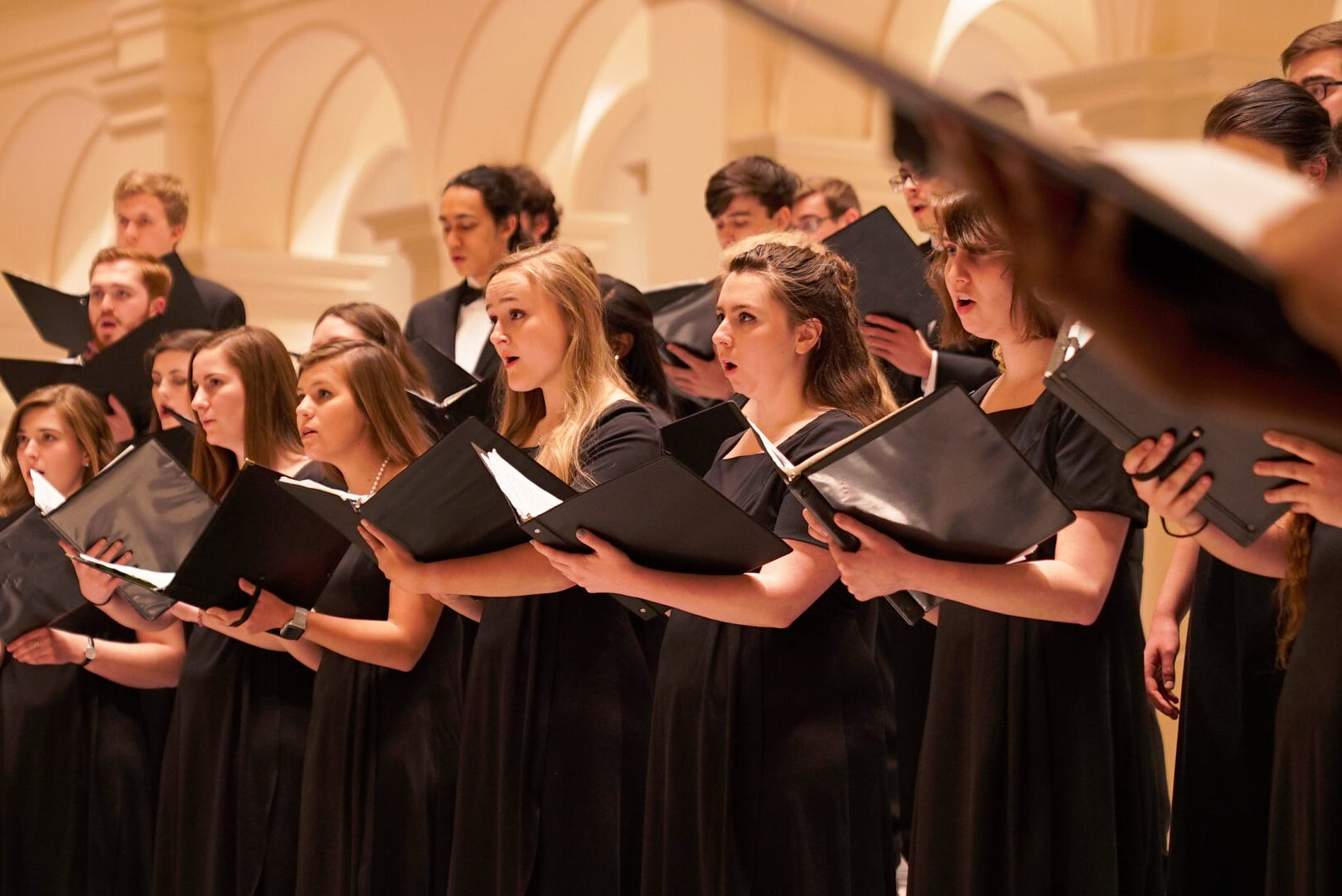 Photograph of the State Chorale performing inside Holy Name of Jesus Cathedral. Image shows a large group of students wearing formal attire, holding folders of sheet music and singing.