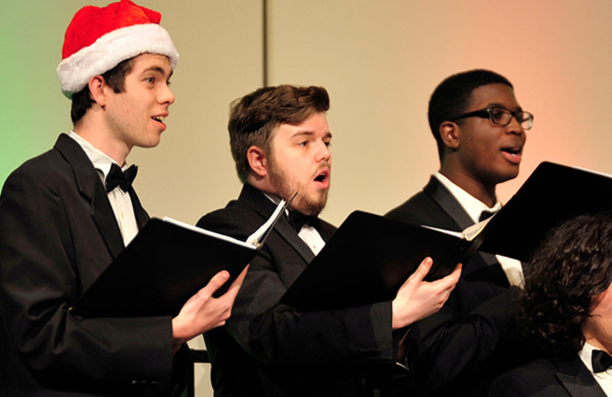 three choir singers perform. The singer on the far right is wearing a Santa hat.